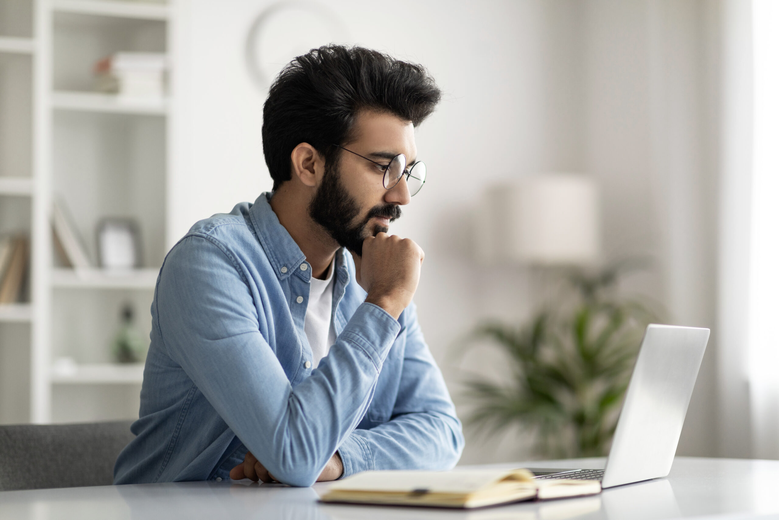 Pensive Young Indian Male Freelancer In Eyeglasses Working With Laptop At Home